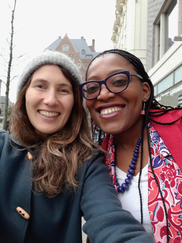 Simone is wearing a grey woollen hat and green coat smiling next to Junnifa wearing a read jacket, necklace and white shirt